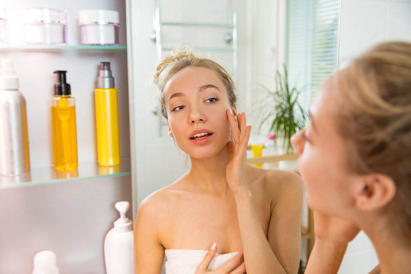 Девушка в полотенце рассматривает себя в зеркало и прикасается к лицу Девушка смотрится в зеркало